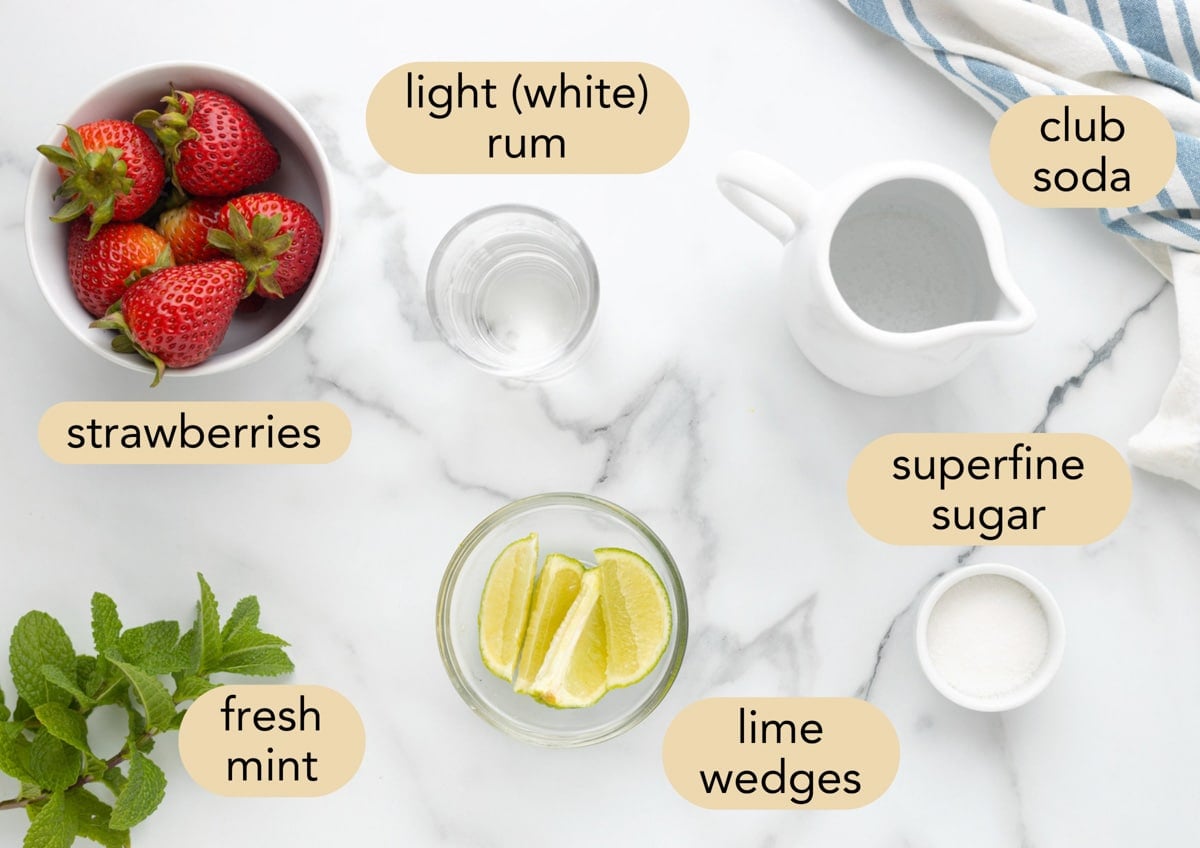 Ingredients for a strawberry mojito arranged on a marble surface: strawberries, fresh mint, lime wedges, light (white) rum, club soda, and superfine sugar.