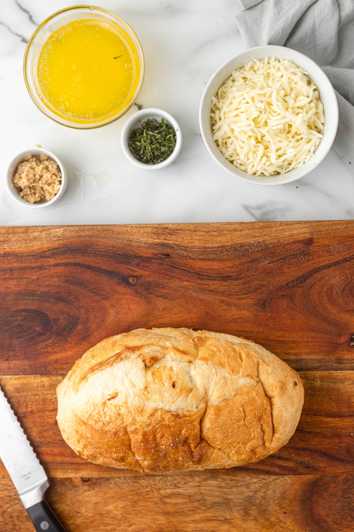 overhead of a loaf of Italian bread on a wood cutting board with bowls of meted butter, shredded mozzarella, chopped herbs, and chopped garlic on white marble