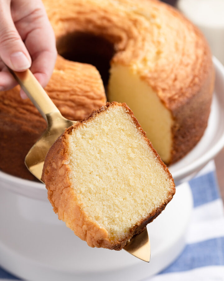slice of vanilla pound cake on a gold cake server in front of a full pound cake on a white cake stand