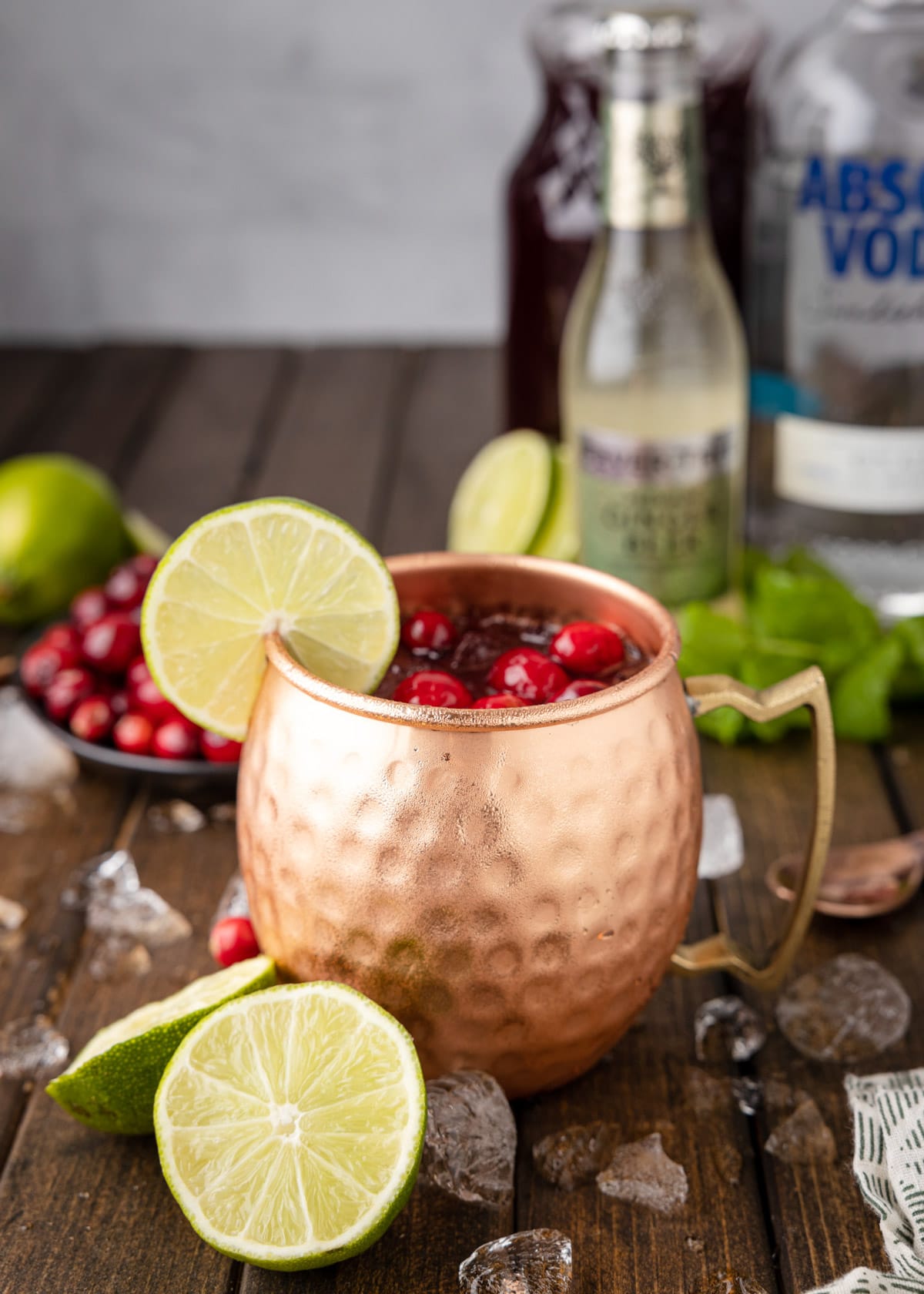 cranberry moscow mule in a copper mug on a wood board, with lime wedges, a bowl of cranberries, and liquor bottles