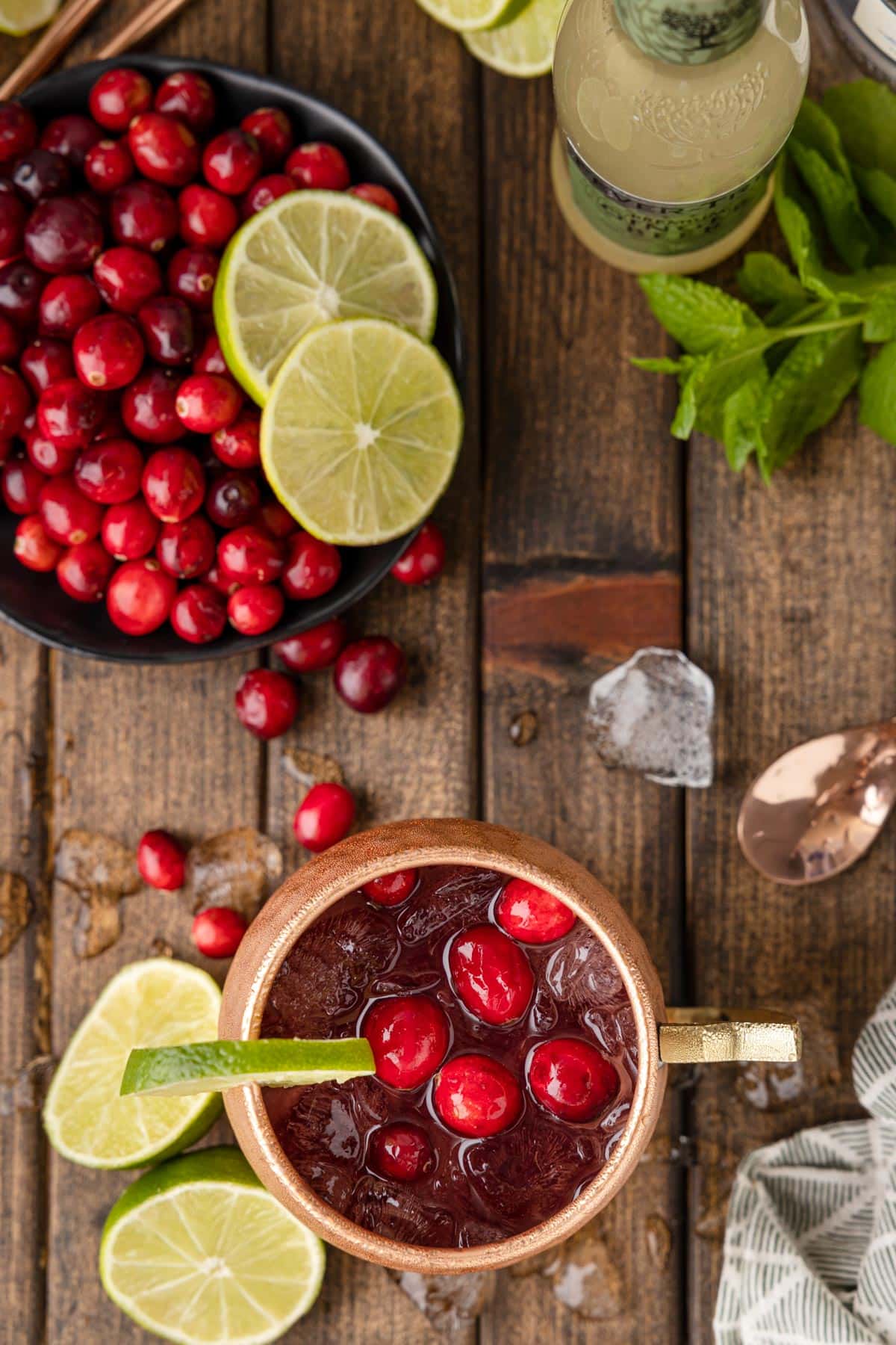 overhead of a garnished Cranberry Moscow Mule on a wooden board surrounded by ice, cranberries, cut lime wedges, and mint sprigs