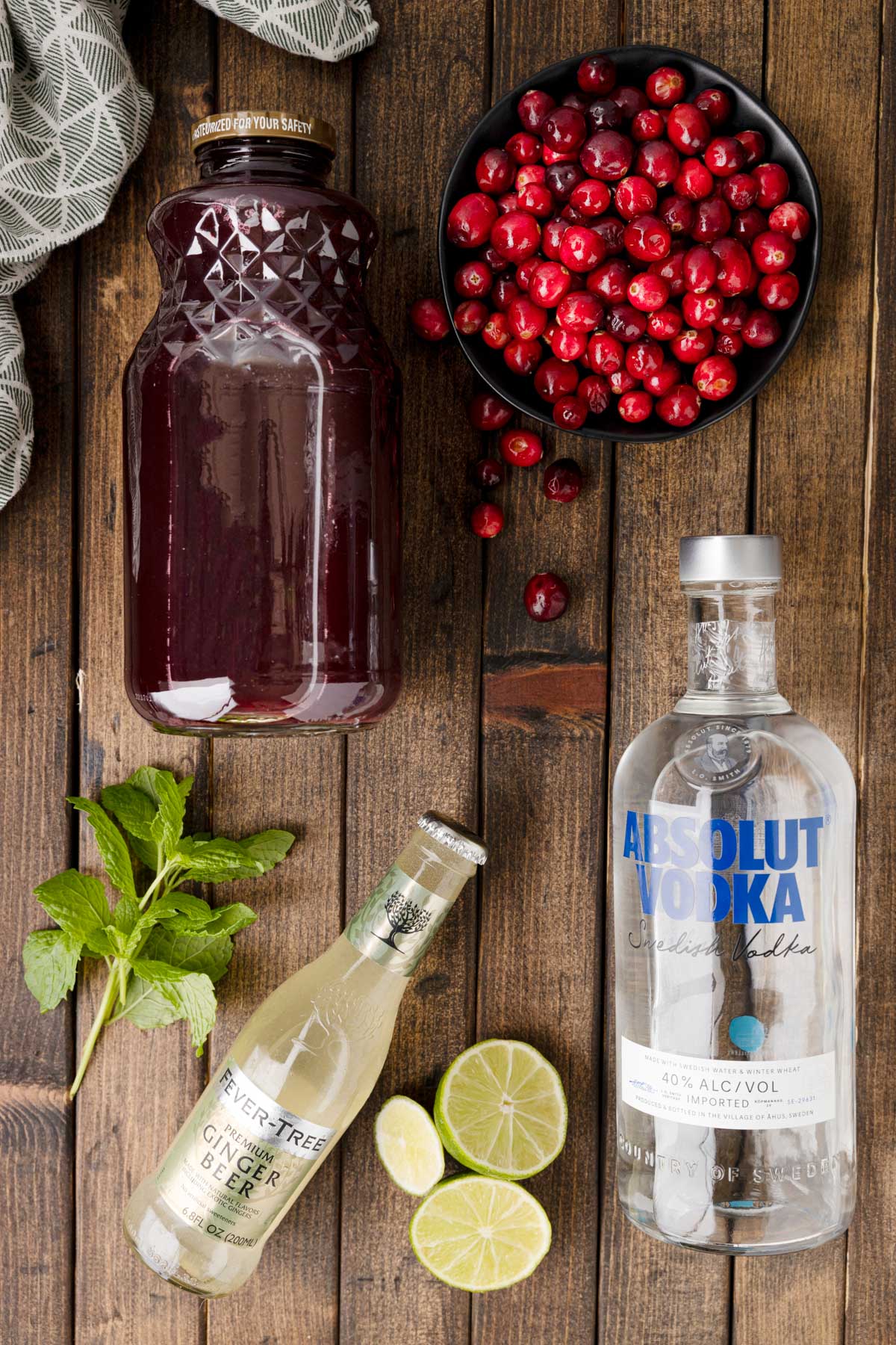 overhead photo of a bottle of cranberry juice, a bottle of Absolut vodka, and a bottle of Fever Tree ginger beer, on a wood board, with cranberries, lime wedges, and fresh mint