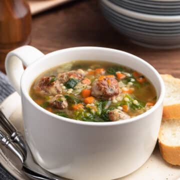 Italian Wedding Soup in a white handled bowl on an ivory ceramic plate with two spoons and slices of Italian bread