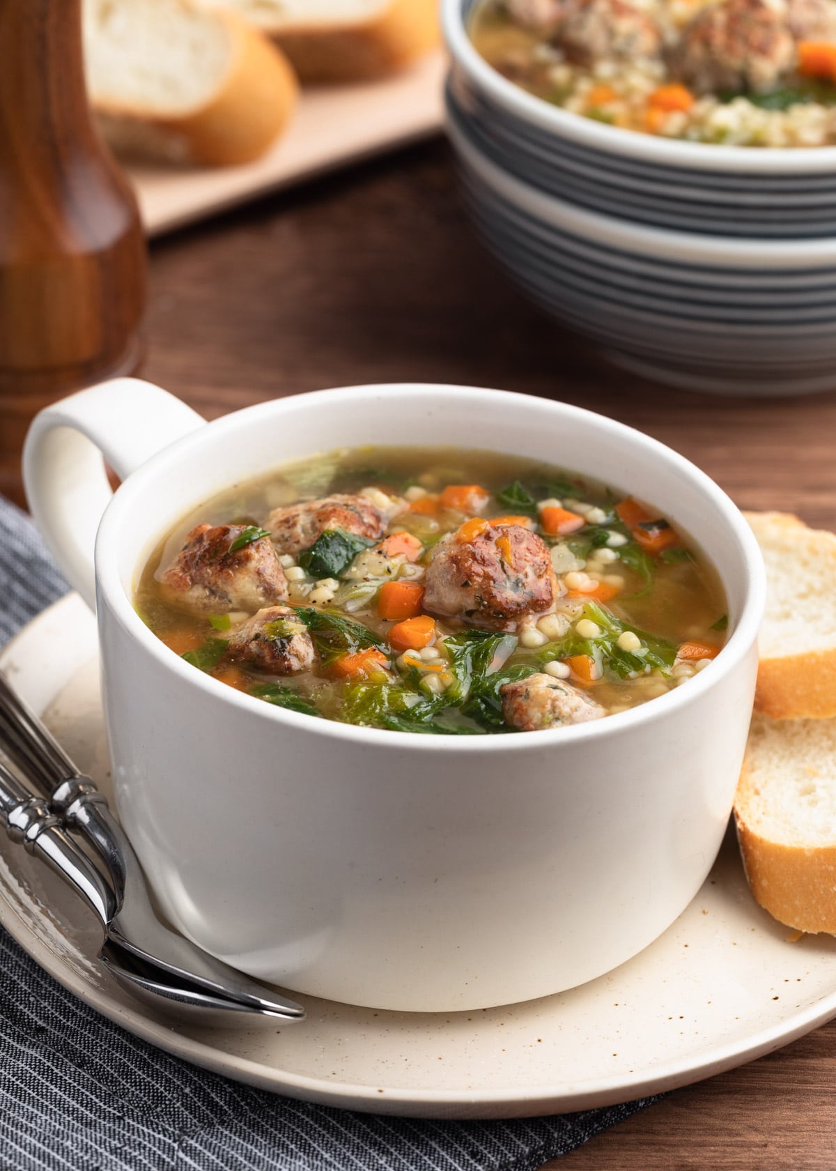 Italian Wedding Soup in a white handled bowl on an ivory ceramic plate with two spoons and slices of Italian bread