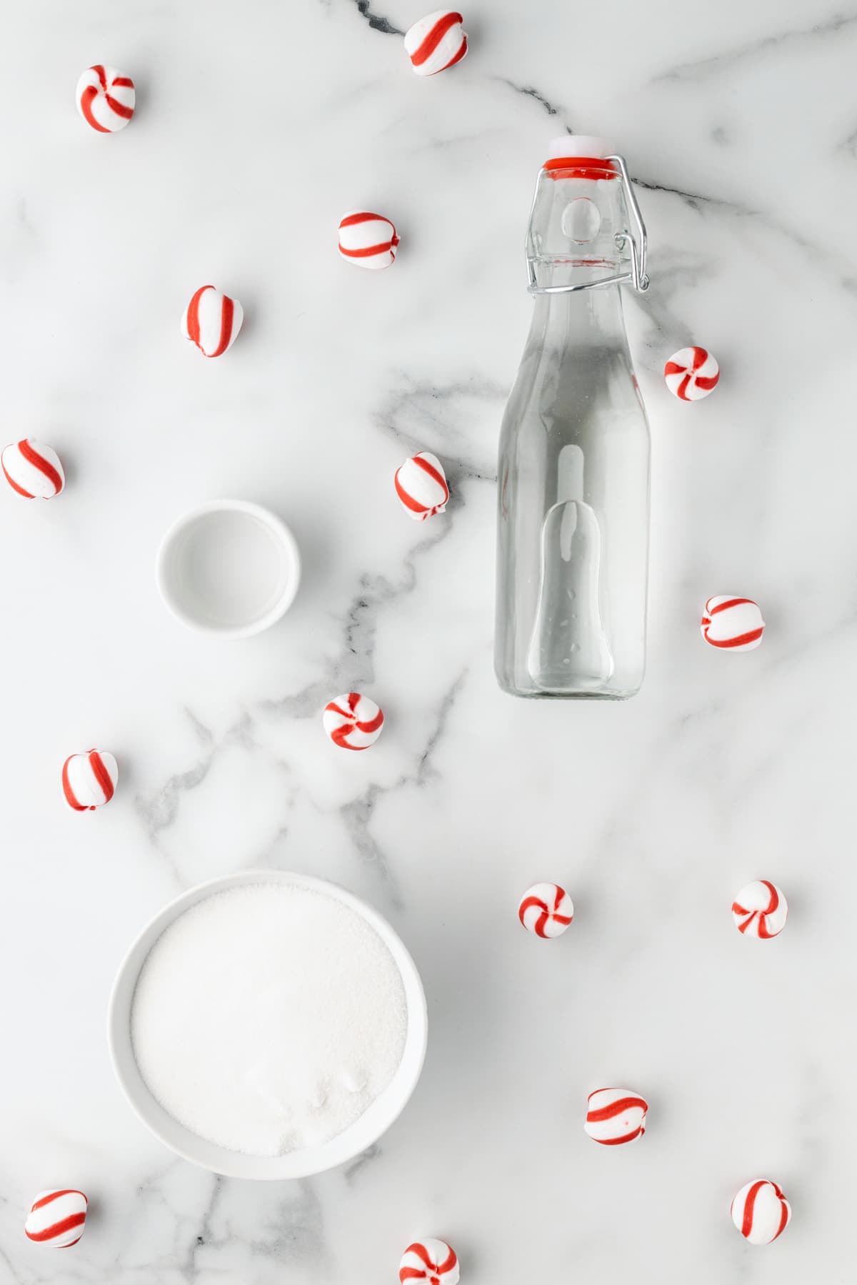 overhead photo of a glass bottle of water, a bowl of granulated sugar, and a small bowl of peppermint extract on a marble background, with scattered peppermint hard candies