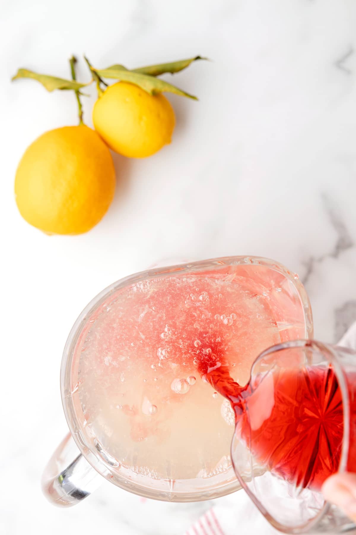 pouring cranberry juice into a pitcher of lemonade