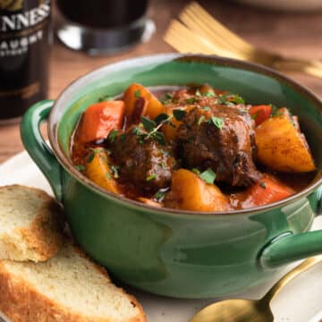 Guinness Beef Stew in a green handled bowl on top of an ivory plate with slices of Irish Soda Bread and a gold tone spoon