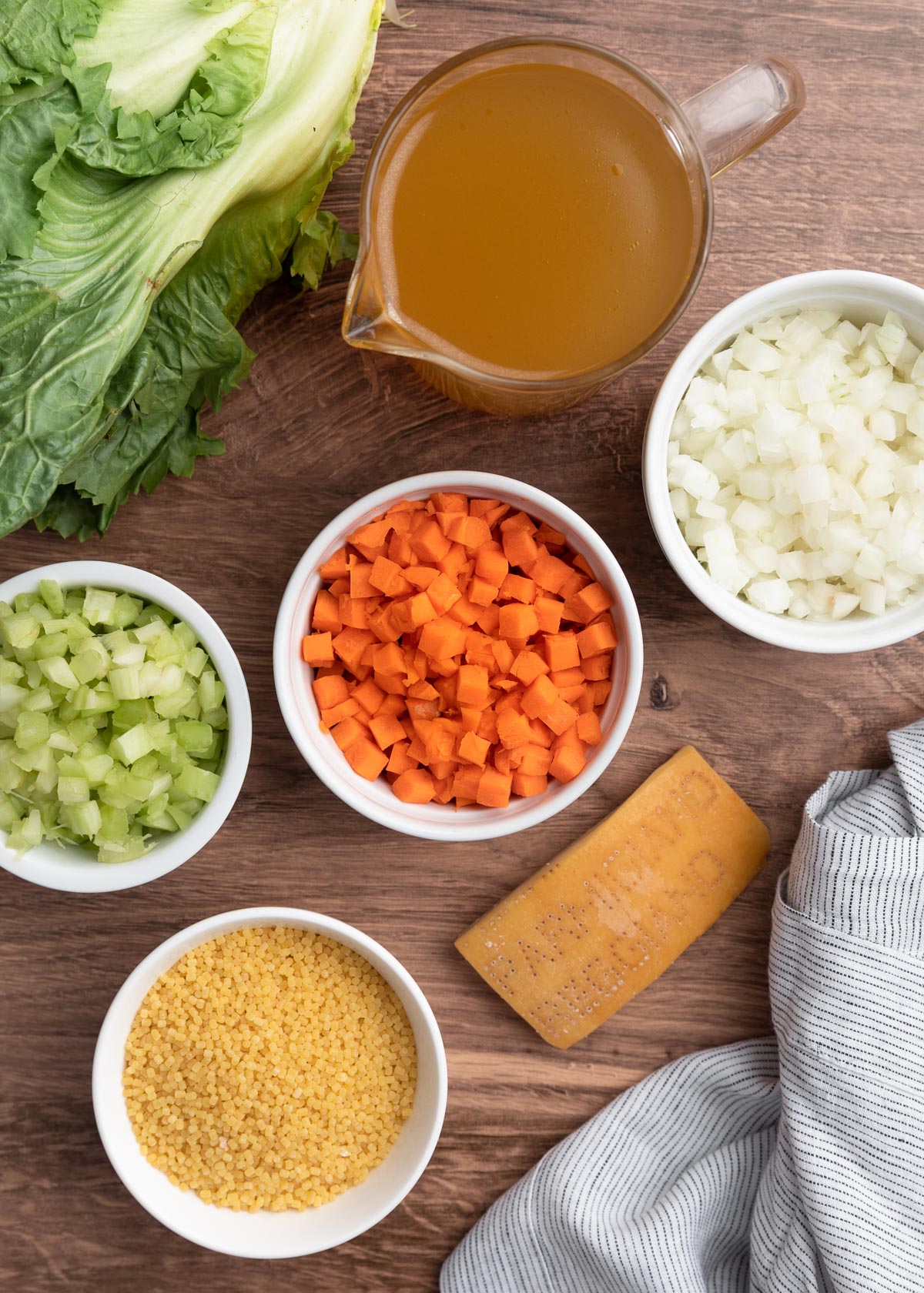 overhead photo of diced carrots, celery, and onions in white bowls, a parmesan cheese rind, a liquid measuring cup filled with chicken broth, and a head of escarole
