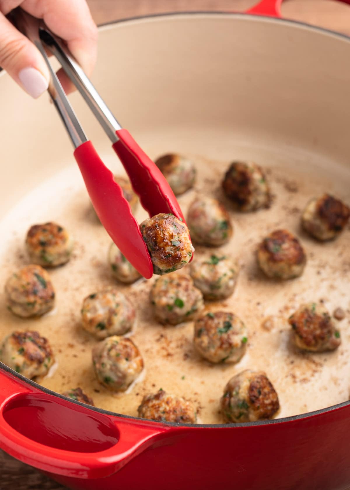 removing a seared beef and pork meatball from a pot with a pair of red tongs