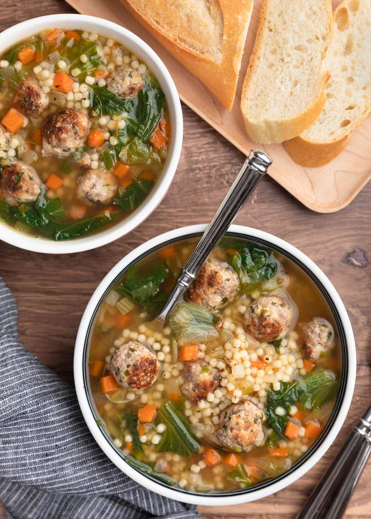 overhead photo of two bowls of Italian Wedding Soup on a wood background with a sliced loaf of Italian bread in the upper right corner