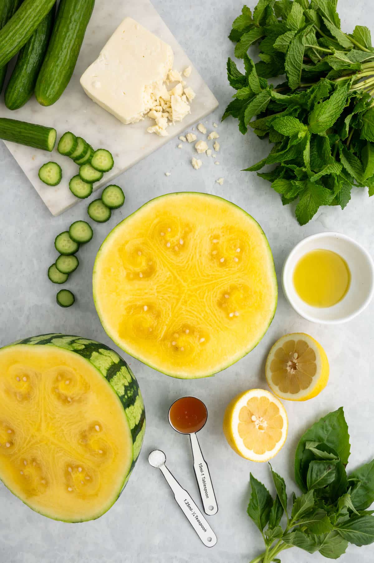 overhead photo of a halved yellow watermelon, lemon halves, sliced persian cucumbers, crumbled feta, honey, olive oil, and herbs on a gray background