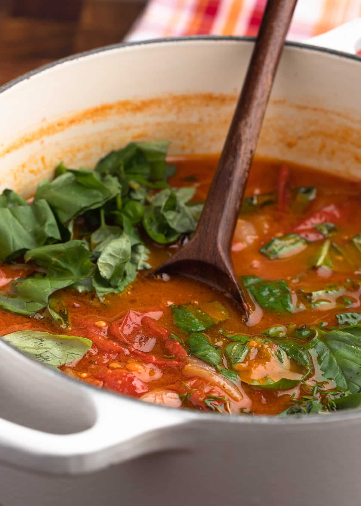 wooden spoon stirring fresh basil leaves into a pot of roasted tomato soup