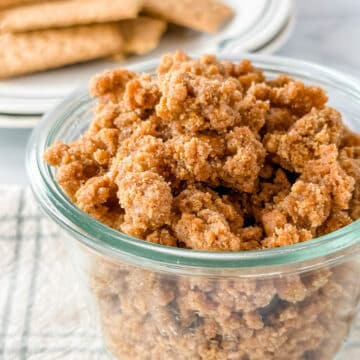 graham cracker crumbles in a glass jar on a white and green check napkin with a plate of graham crackers in the background