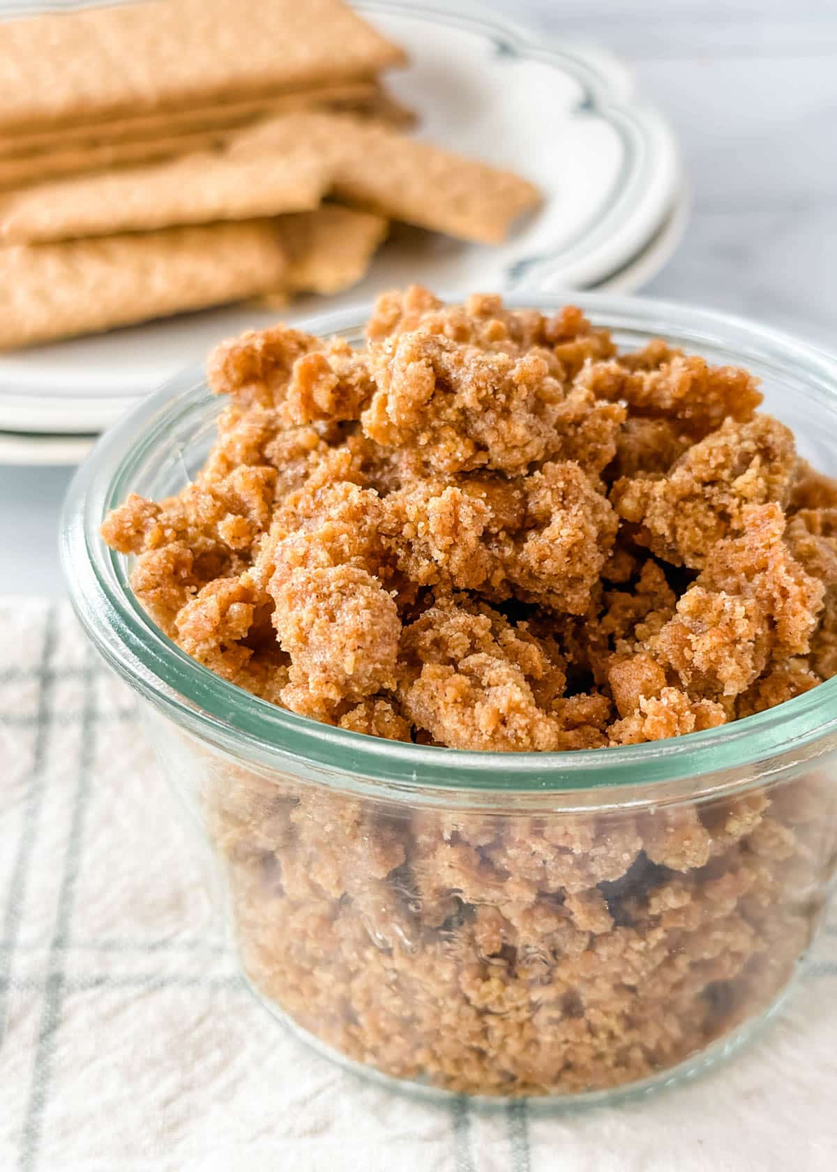 graham cracker crumbles in a glass jar on a white and green check napkin with a plate of graham crackers in the background