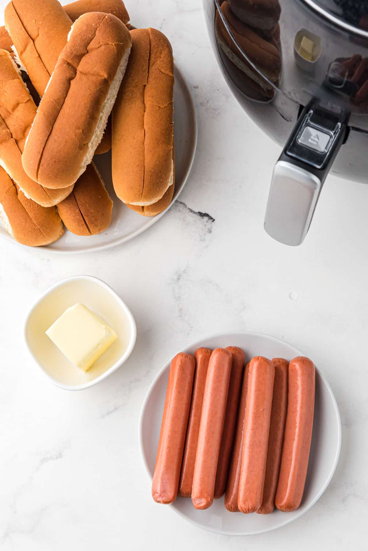 overhead photo of hot dog buns, uncooked hot dogs, butter, and an air fryer on white marble