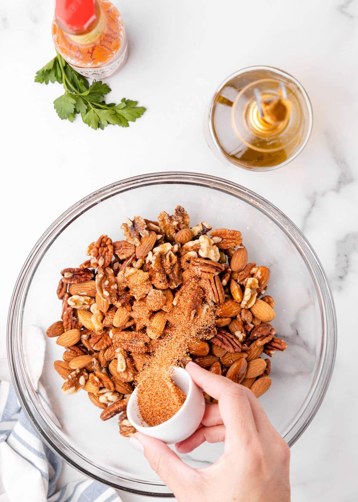 overhead photo showing cajun seasoning being poured into a glass bowl of mixed nuts on a marble board