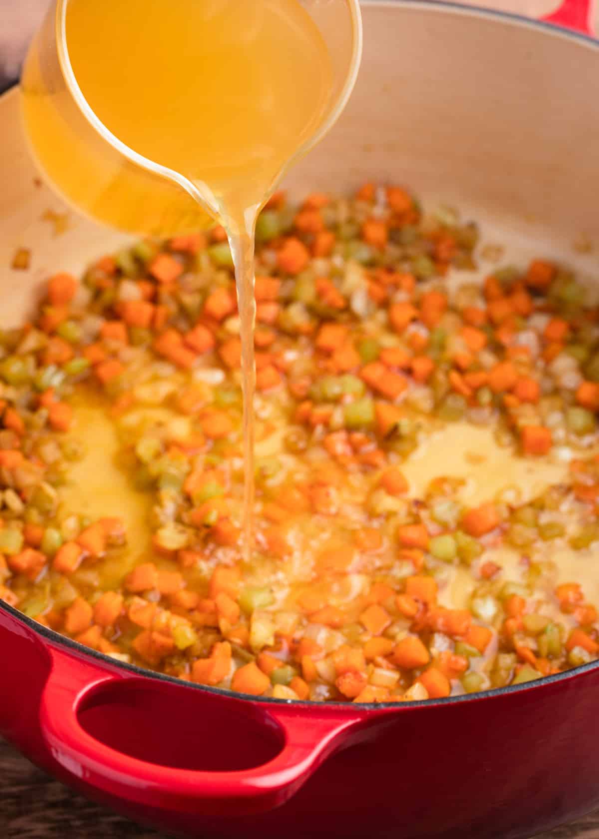 pouring chicken stock from a glass liquid measuring cup into a red enameled cast iron dutch oven with sauteed carrots, celery, and onions