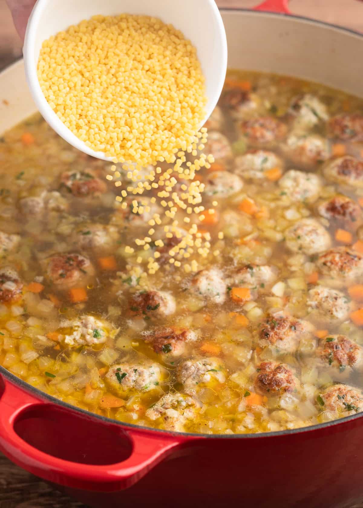 acini di pepe pasta being poured into a pot of Italian Wedding Soup from a white ceramic bowl