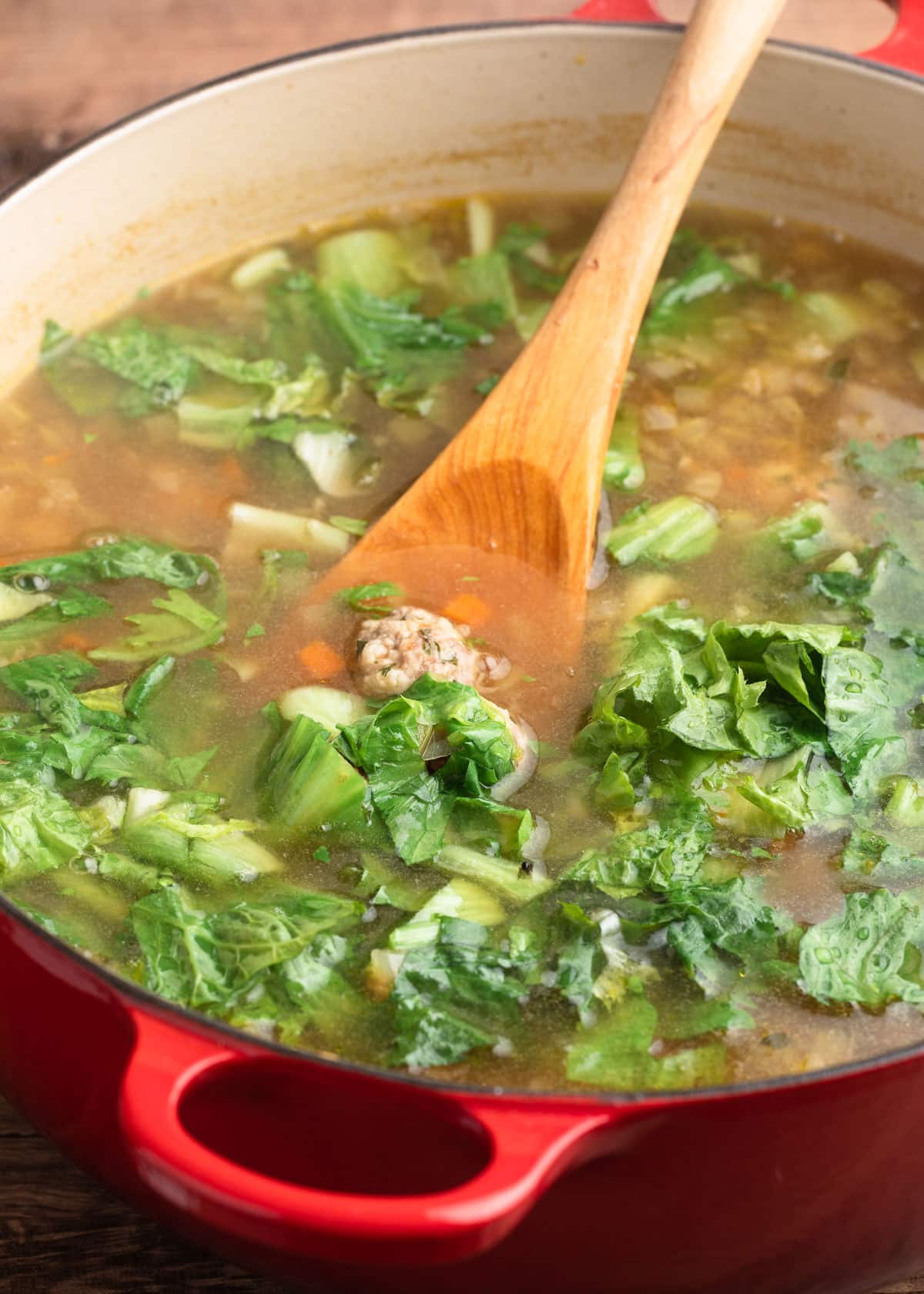 chopped escarole being stirred into a pot of Italian Wedding Soup with a wooden spoon