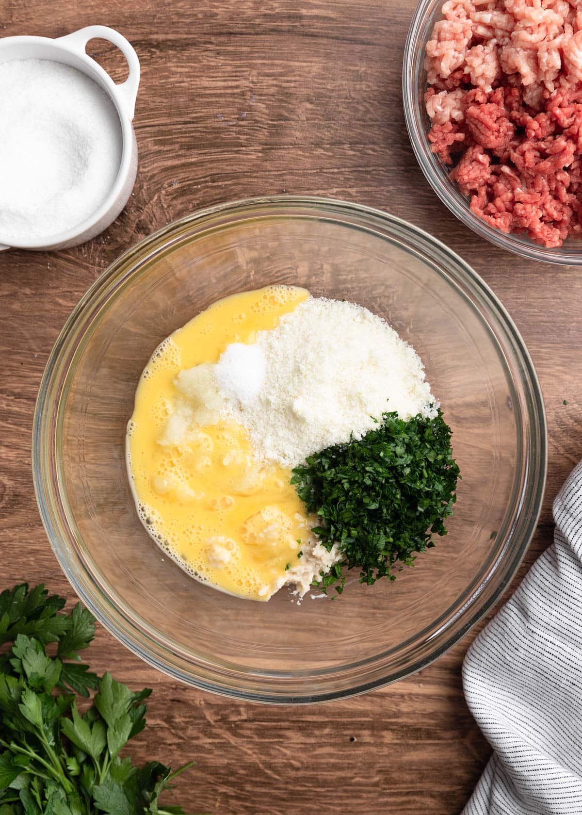 overhead photo of beaten egg, pecorino romano cheese, and chopped parsley in a glass bowl on a wood board
