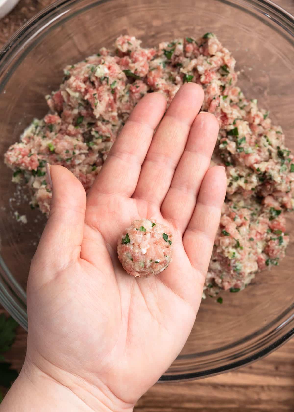 overhead photo of an open hand holding a small beef and pork meatball over a bowl of the unrolled meatball mixture