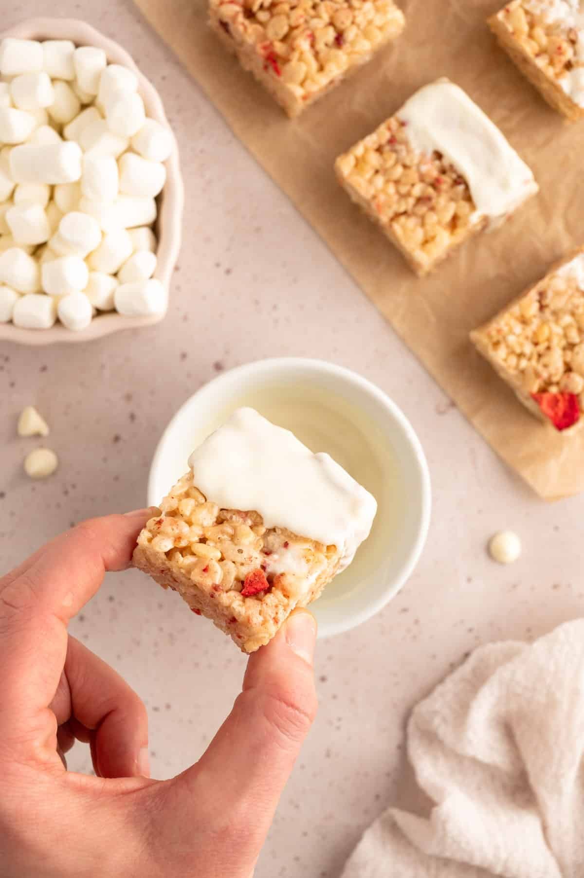 overhead photo of a cut strawberry rice krispie treat being dipped into a bowl of melted white chocolate