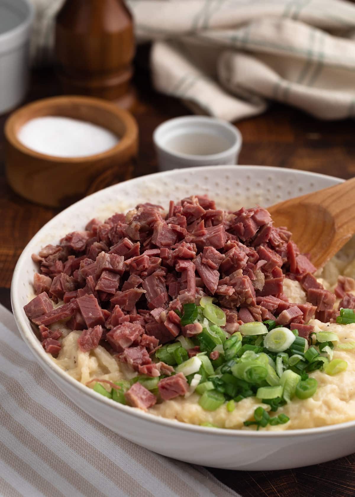 diced corned beef and sliced scallions added to an ivory bowl of potato pancake batter with a wooden spoon