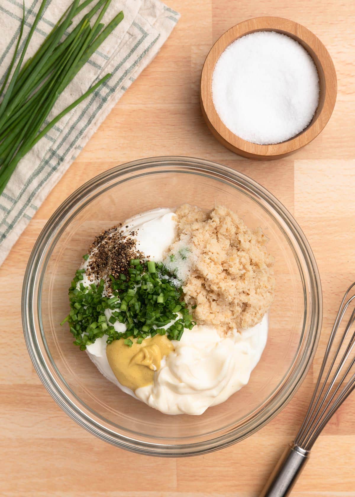 overhead photo of ingredients for creamy horseradish sauce in a mixing bowl, with a whisk and wooden bowl of salt on the side