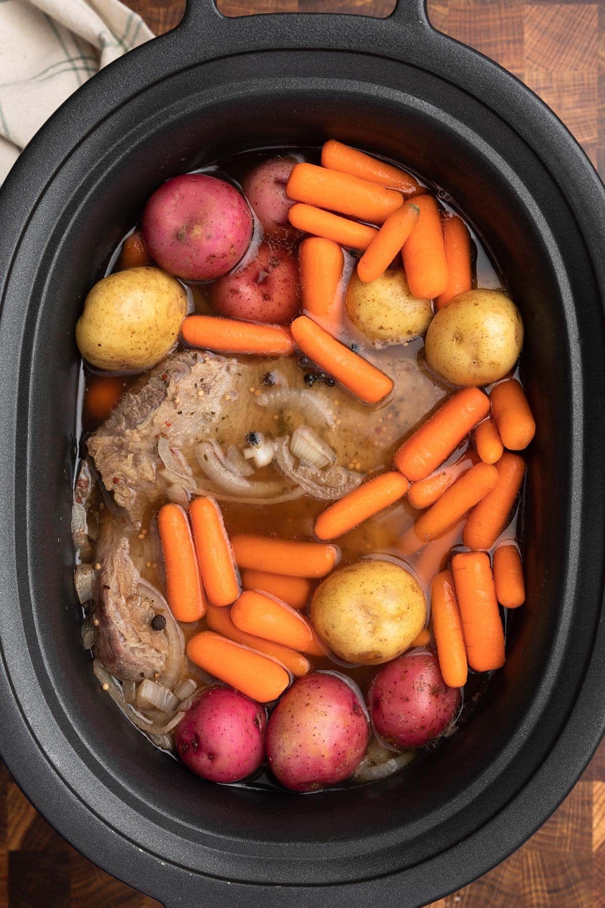 overhead photo of corned beef in a slow cooker insert with raw baby potatoes and carrots