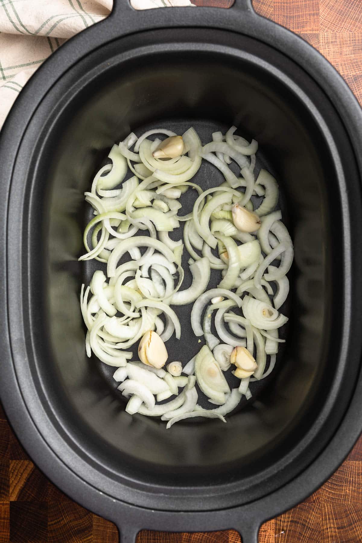 overhead of sliced yellow onions and whole garlic cloves in a black slow cooker insert