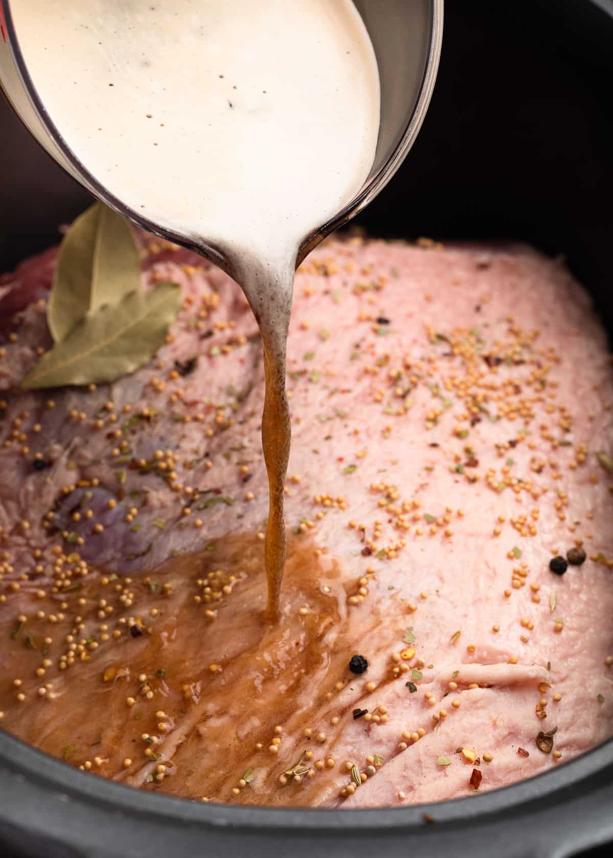 stout beer being poured from a liquid measuring cup over an uncooked corned beef brisket in a slow cooker insert