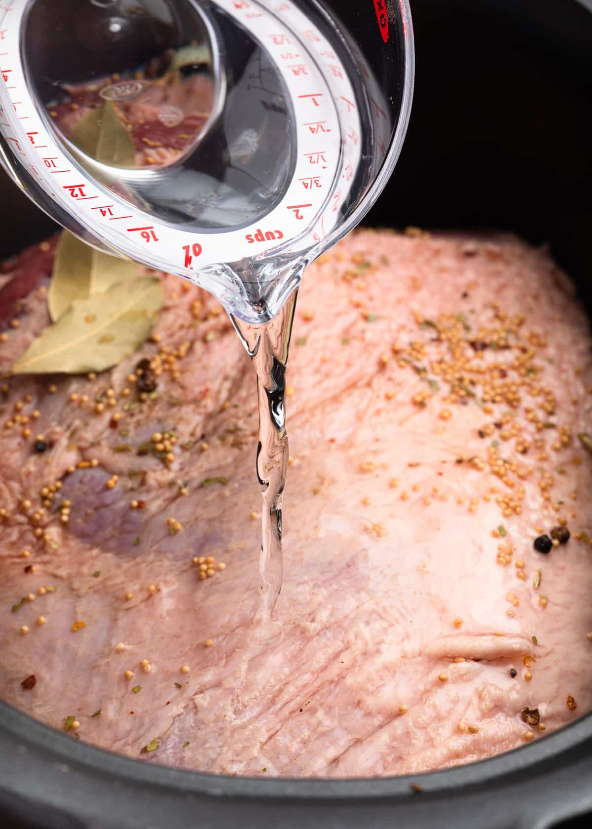 water being poured from a liquid measuring cup over an uncooked corned beef brisket in a slow cooker insert