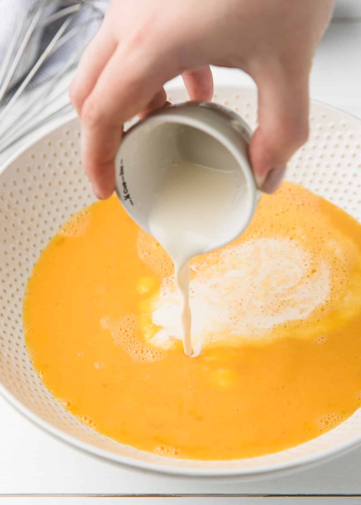 pouring heavy cream into a white ceramic bowl of beaten eggs