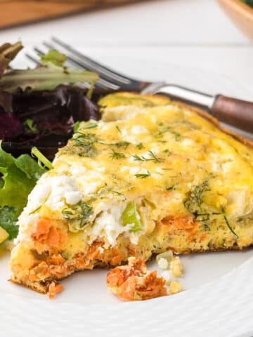 wedge of smoked salmon and goat cheese frittata on a white plate with spring greens, a lemon wedge, and a wood and stainless steel fork