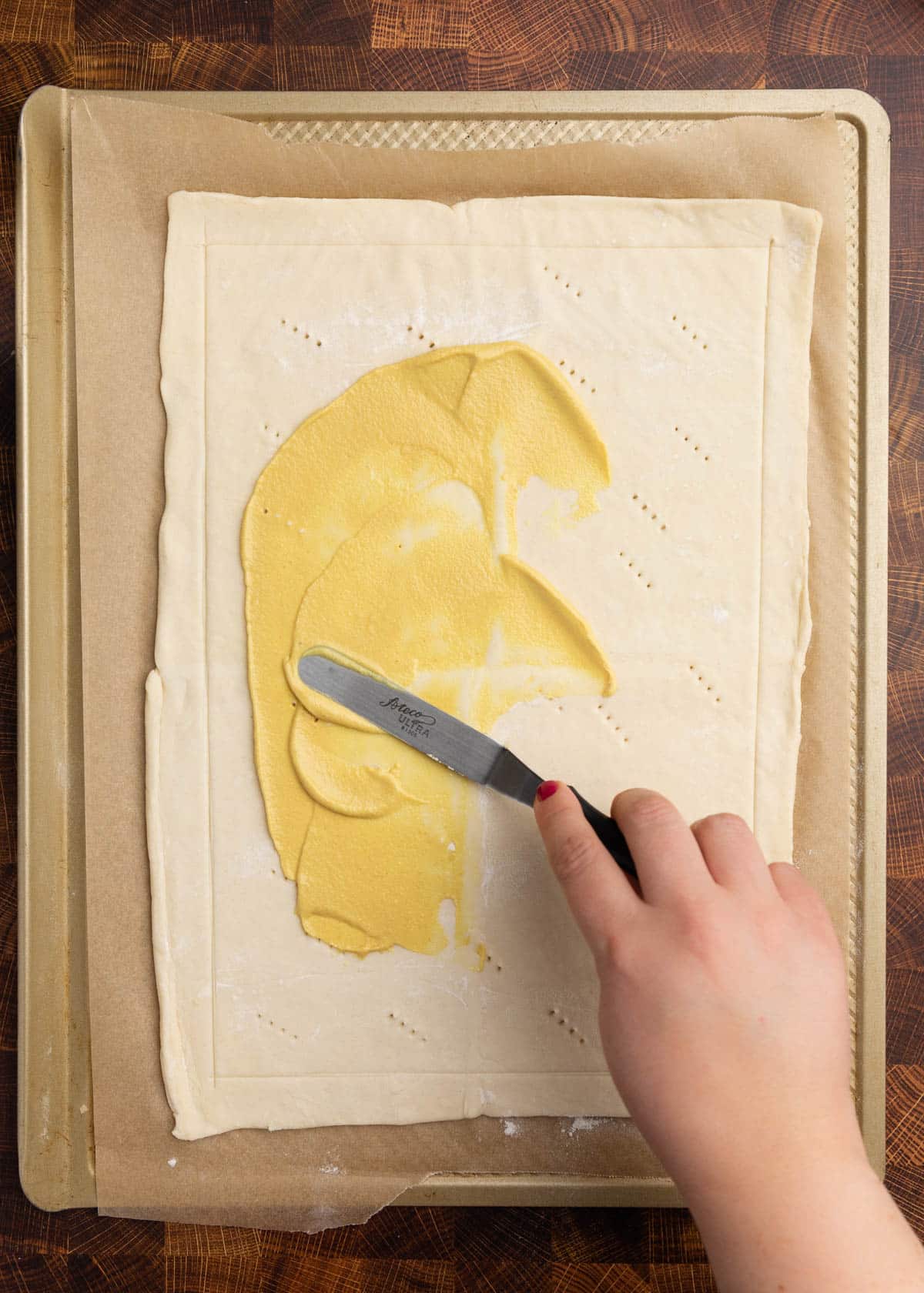 overhead photo of spreading Dijon mustard onto a puff pastry dough on a parchment lined baking sheet
