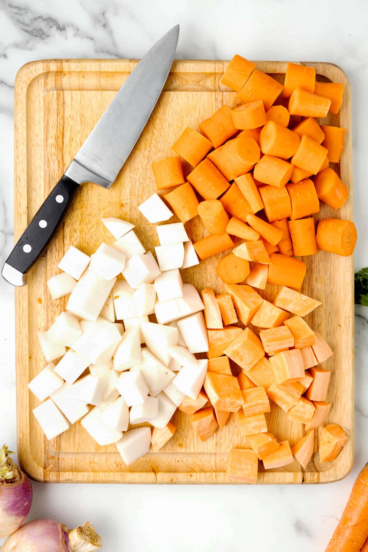 overhead photo of diced carrots, sweet potatoes, and turnips on a wooden cutting board with a chef's knife