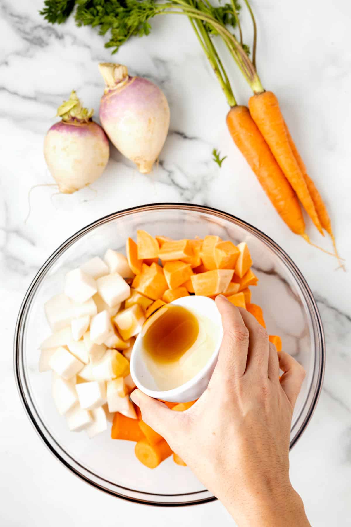 overhead photo of cubed root vegetables in a glass bowl with maple syrup being poured over the top
