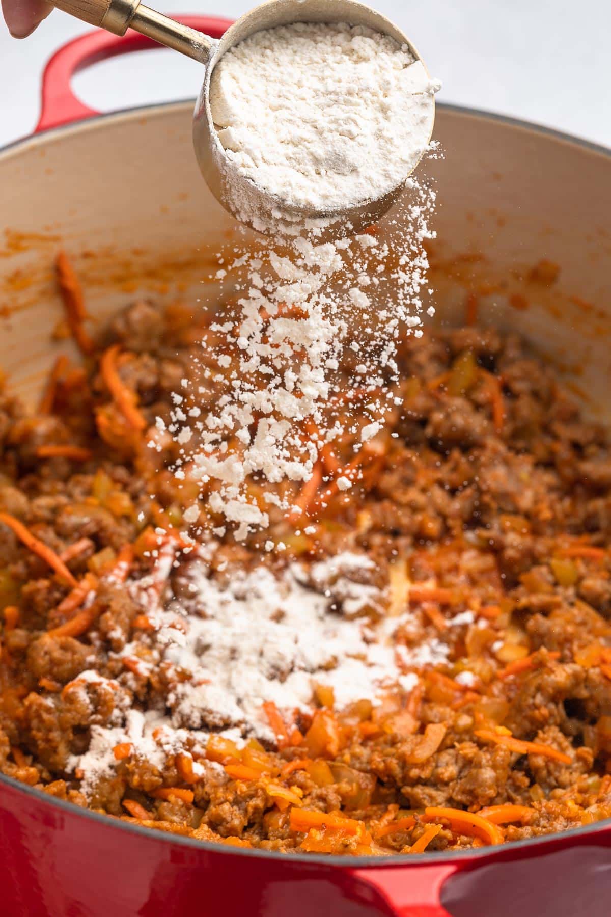 flour being sprinkled into brown sausage and sauteed aromatics in a red enameled cast iron Dutch oven