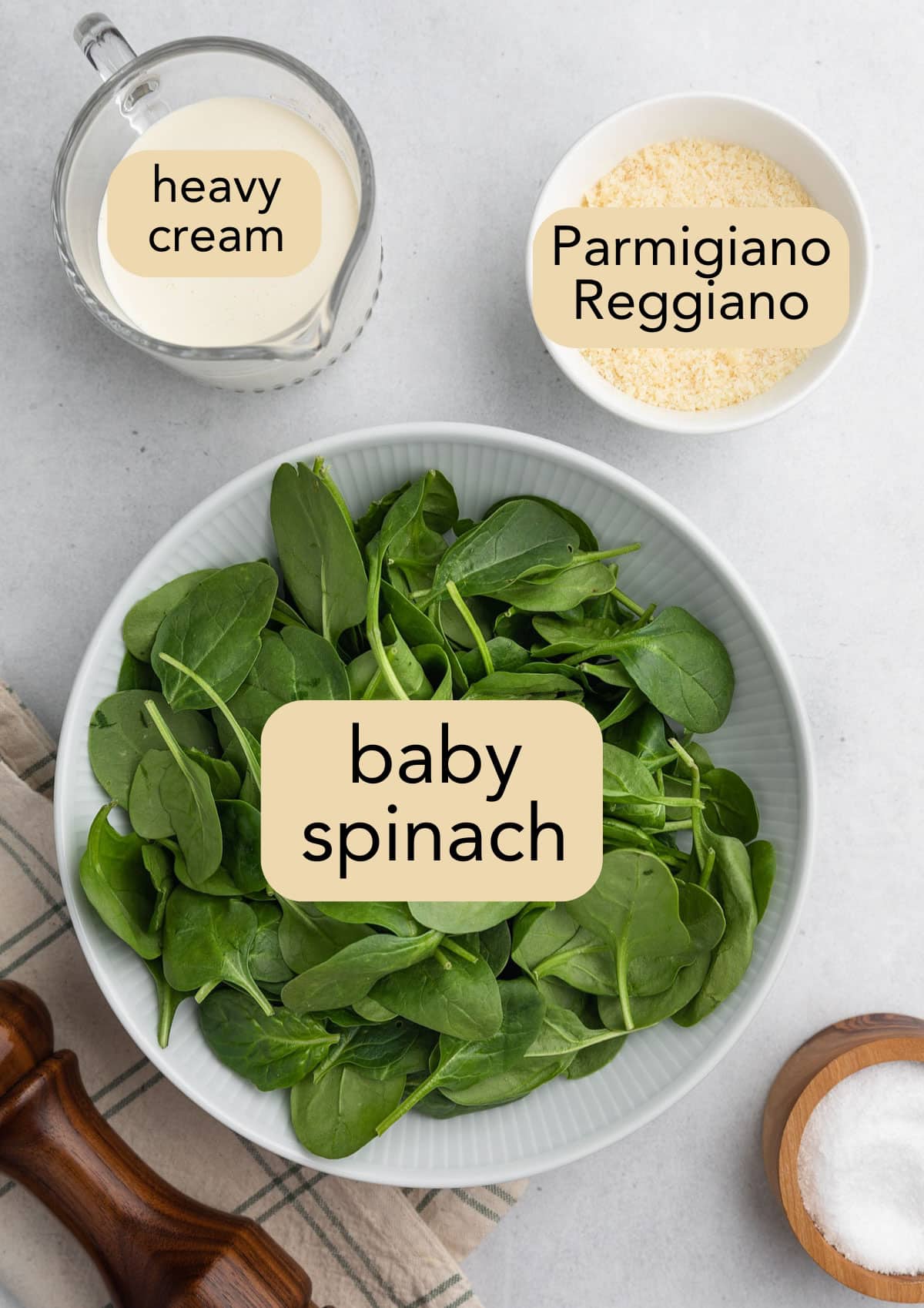 overhead photo of a bowl of baby spinach, a cruet of heavy cream, a bowl of grated parmesan cheese, and a bowl of salt on a white background with text label overlays