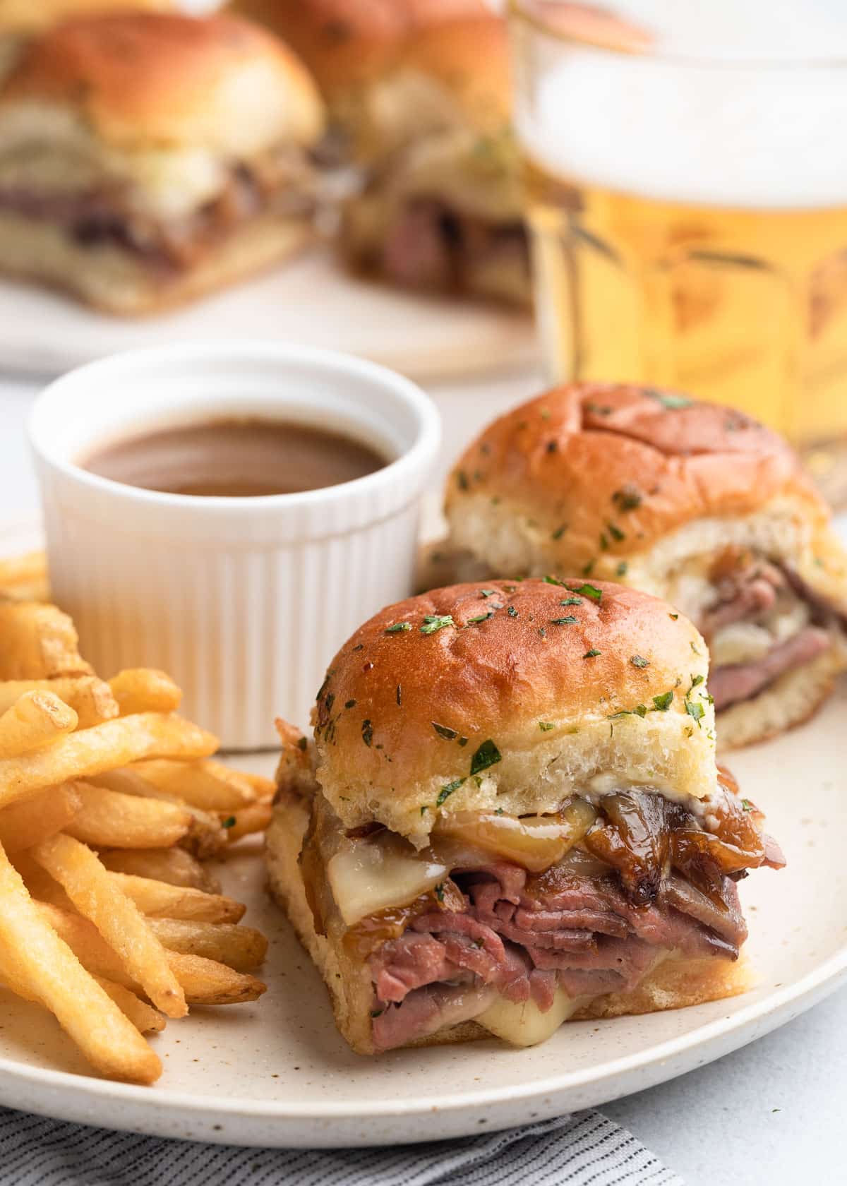 french dip sliders on an ivory ceramic plate with french fries and a cup of au jus