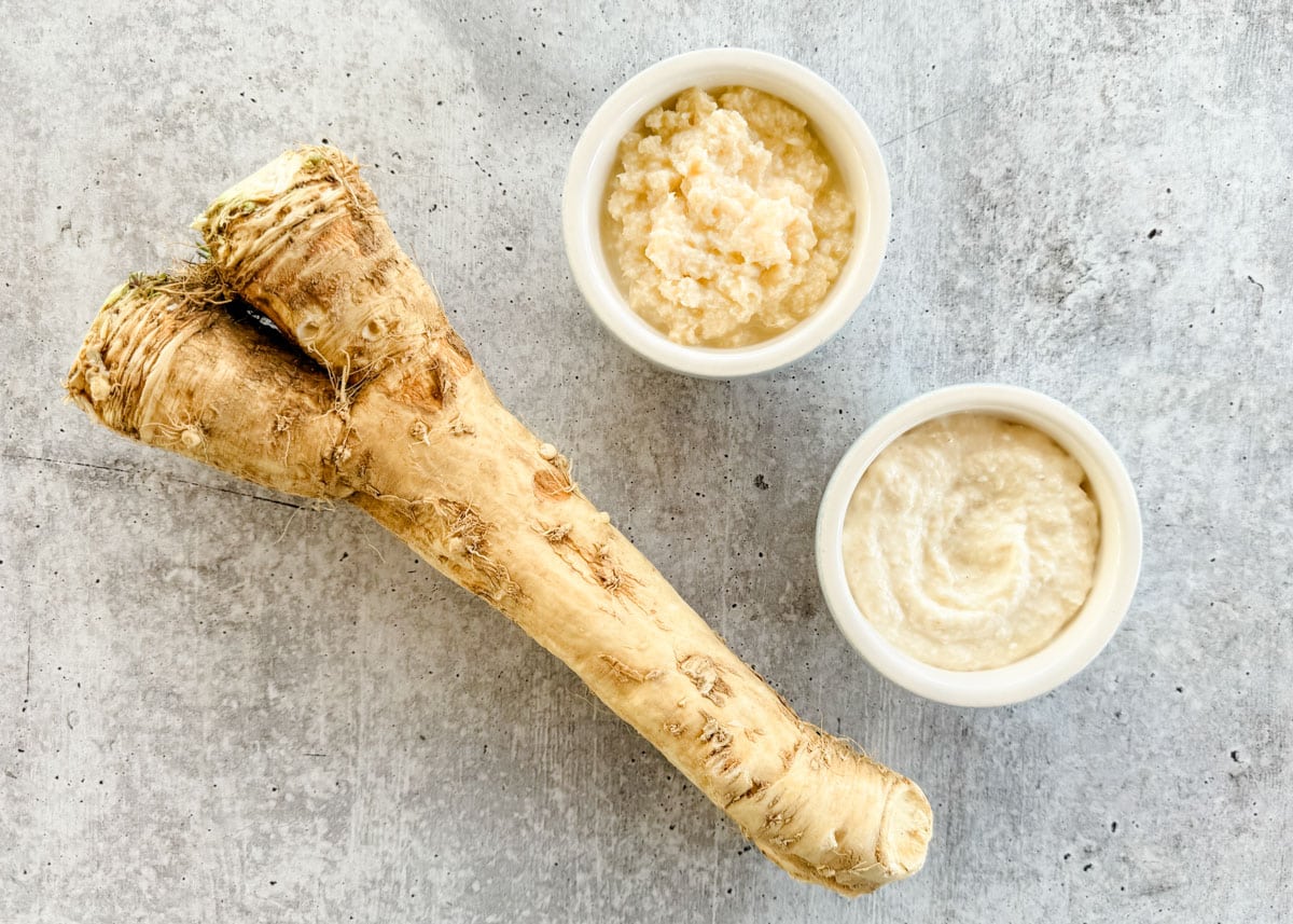 horseradish root, a bowl of prepared horseradish, and a bowl of horseradish sauce on a gray board