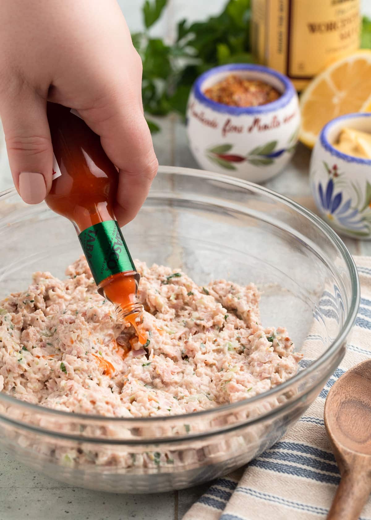 shaking hot sauce from a small glass bottle into a glass bowl of Deviled Ham spread