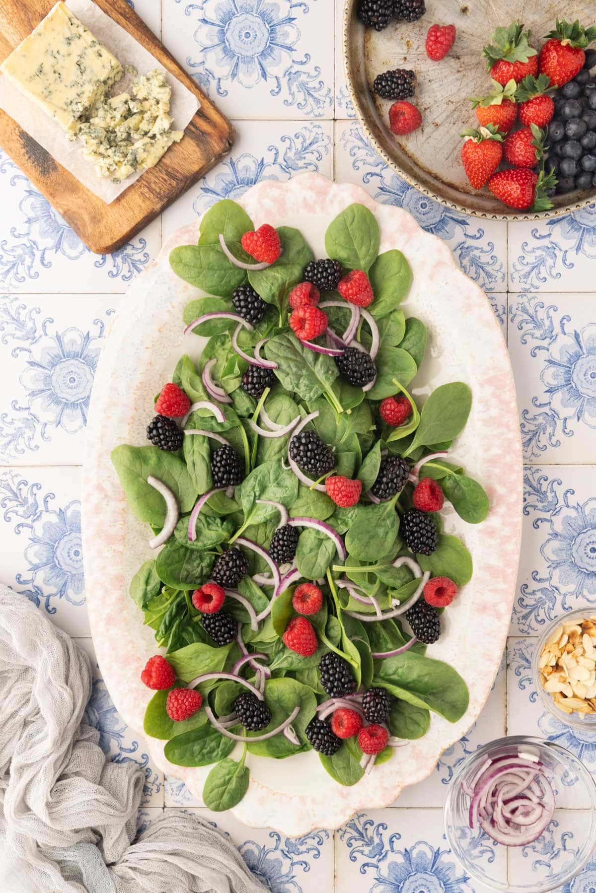 overhead of baby spinach, red onions, blackberries, and raspberries on an oval ivory platter sitting on a blue and white tile background.