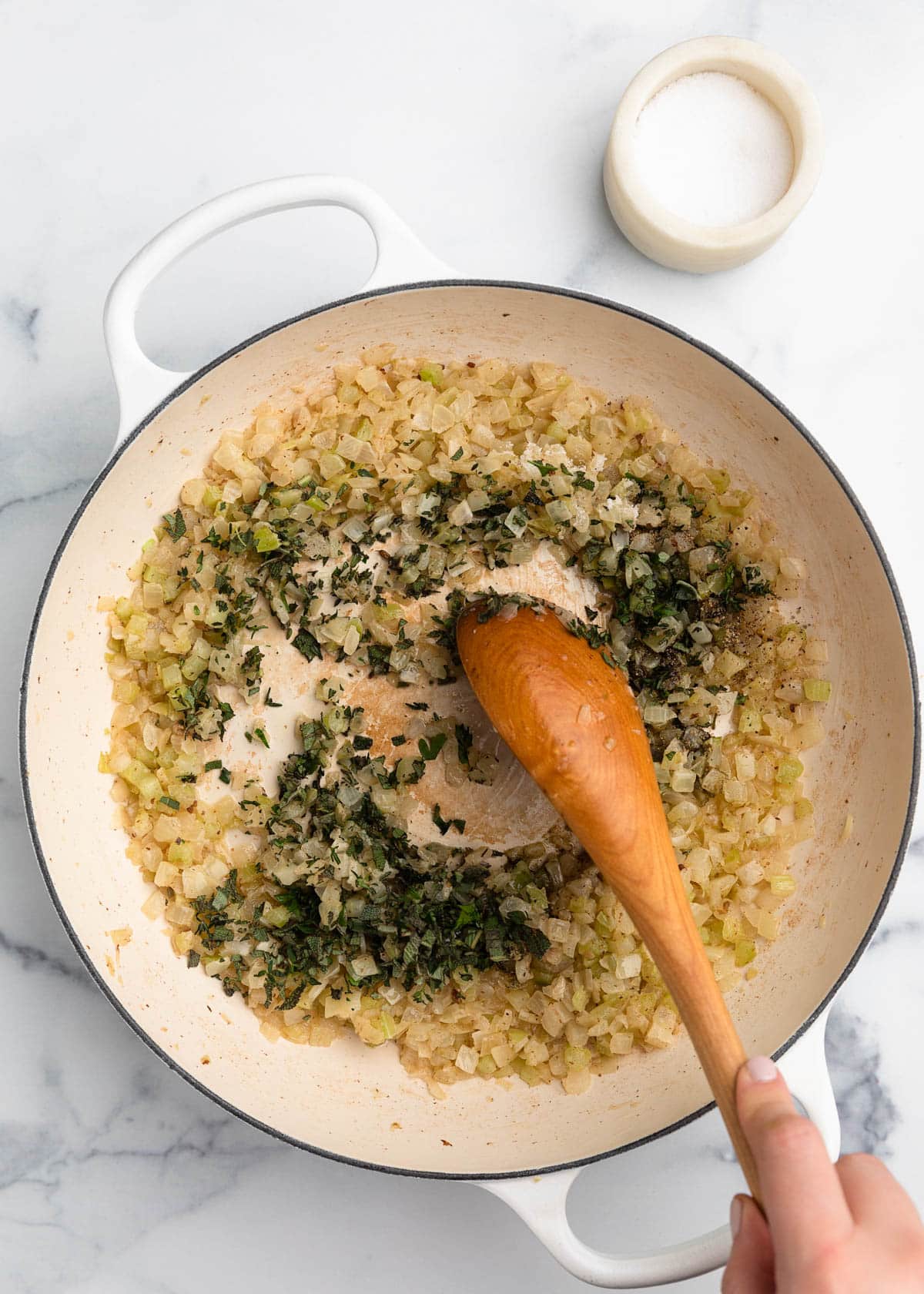 fresh chopped herbs and seasonings being stirred into cooked celery and onions in a white enameled cast iron dutch oven with a wooden spoon