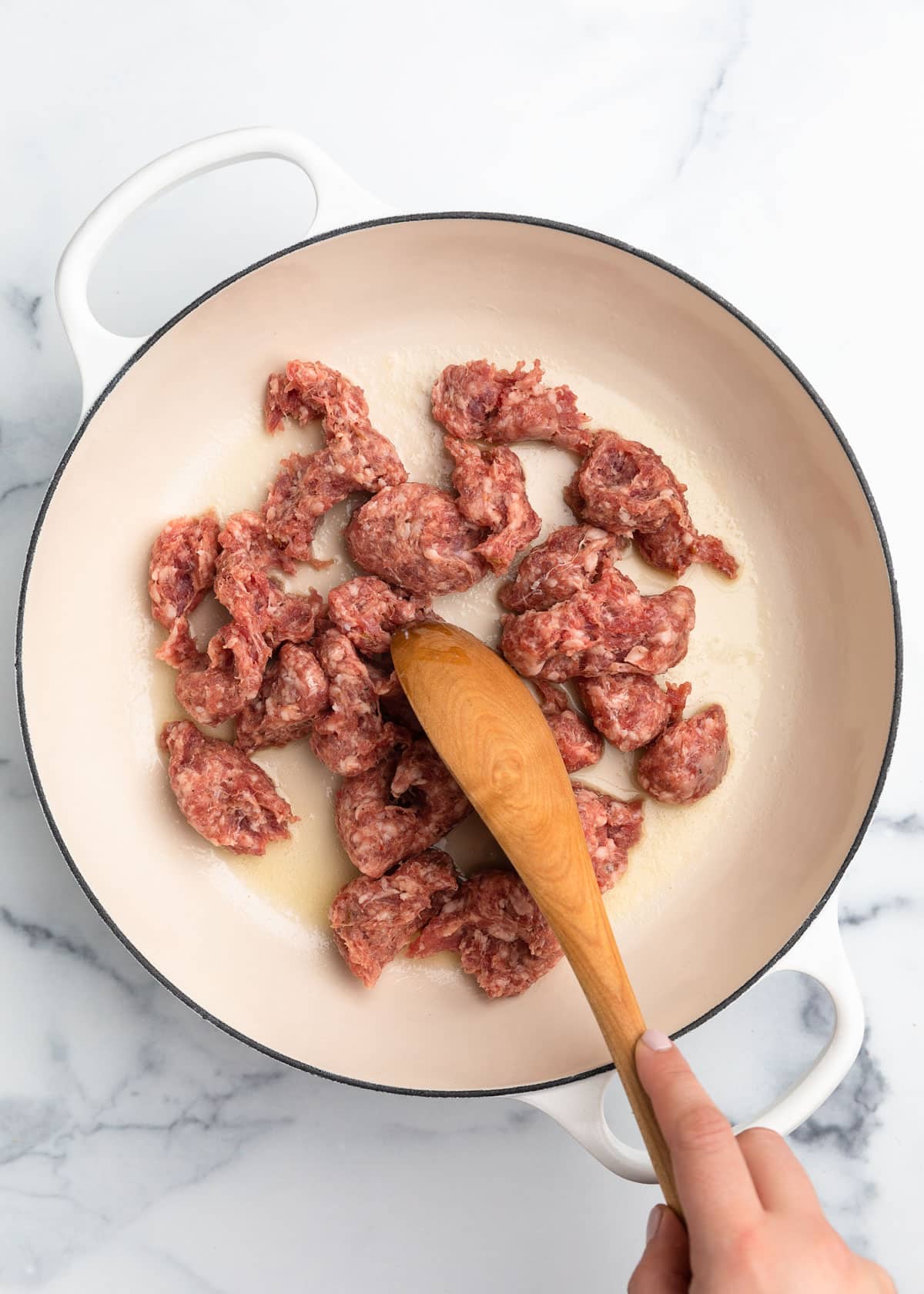 raw loose sausage meat in a white enameled cast iron dutch oven with a wooden spoon