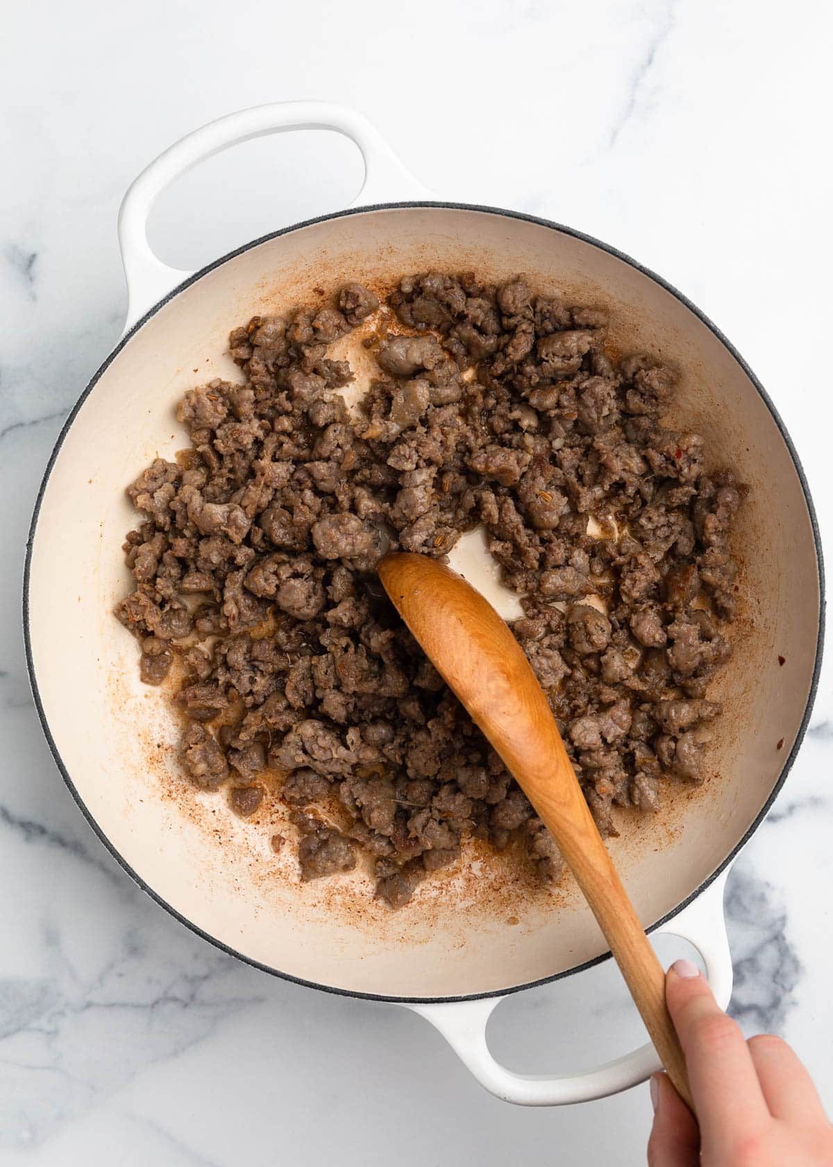 browned Italian sausage crumbles in a white enameled cast iron dutch oven with a wooden spoon