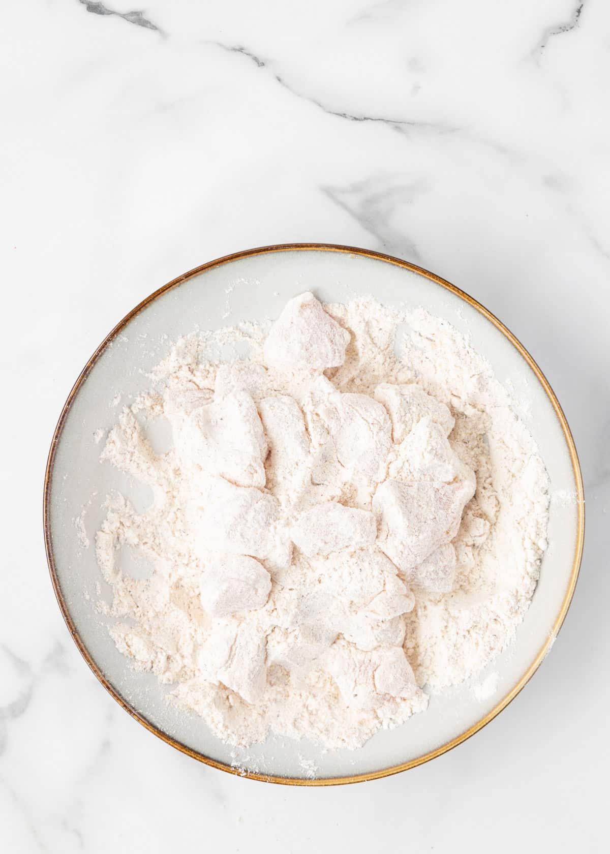 raw chicken cubes dredged in seasoned flour in a shallow bowl on a white marble board