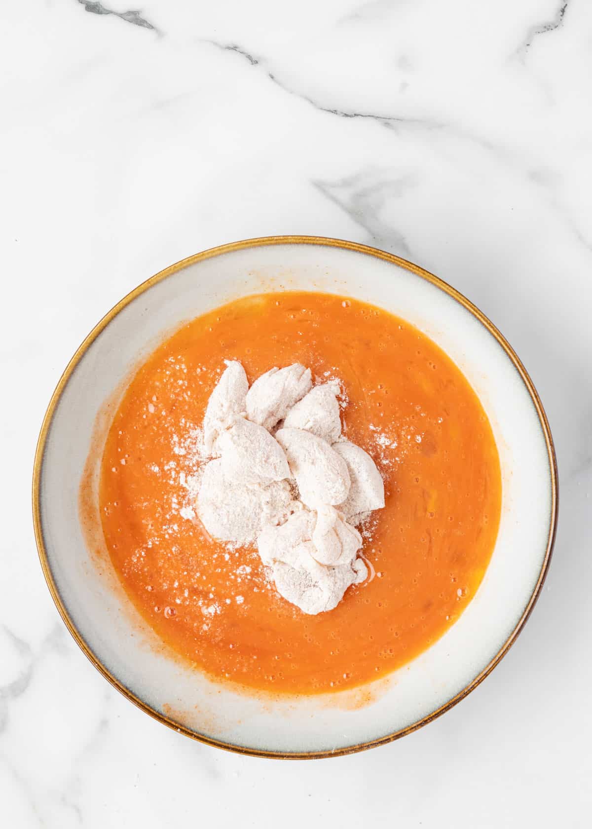chicken tender pieces dredged in flour in a buffalo sauce egg wash in a shallow bowl on a white marble board