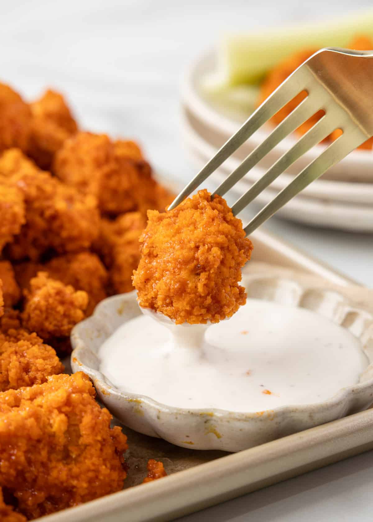 air fryer buffalo chicken bite being dipped into a bowl of ranch dressing from a fork. A pile of chicken bites surrounds the bowl on a rimmed baking sheet.