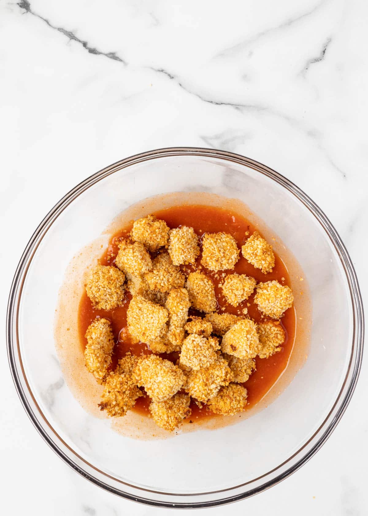 air fried buffalo chicken bites sitting on top of buffalo sauce, untossed, in a glass bowl on a white marble board